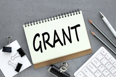 Grants.,Text,On,Open,Notebook,Near,White,Keyboard,And,Pencils