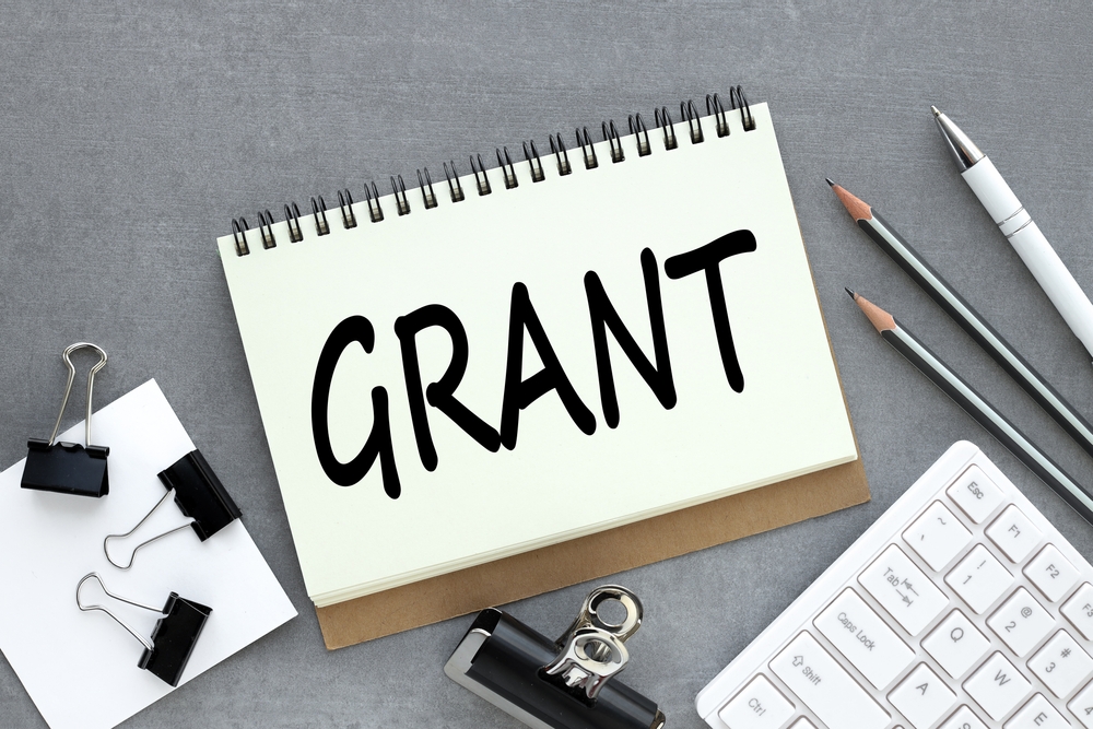 Grants.,Text,On,Open,Notebook,Near,White,Keyboard,And,Pencils